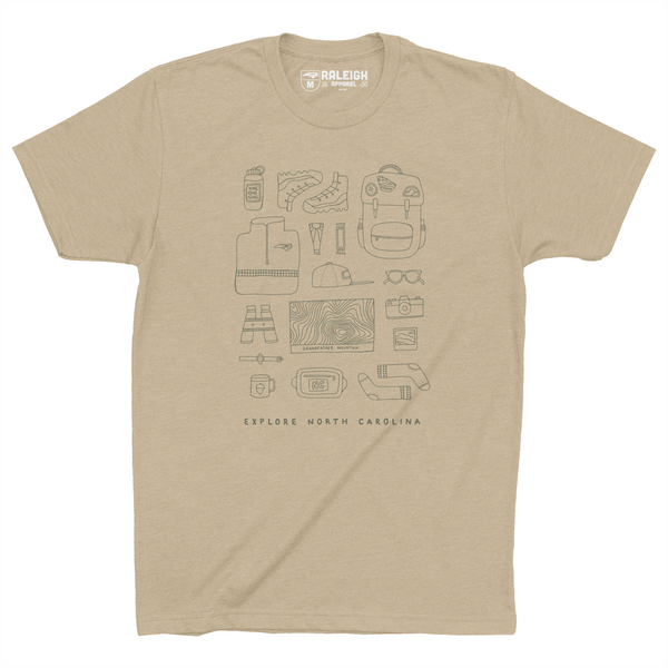 Cream colored t-shirt with different hiking accessories such as boots, backpack, topo map, binoculars, sunglasses and more, drawn in black print.  Bottom of the shirt says Explore North Carolina. 